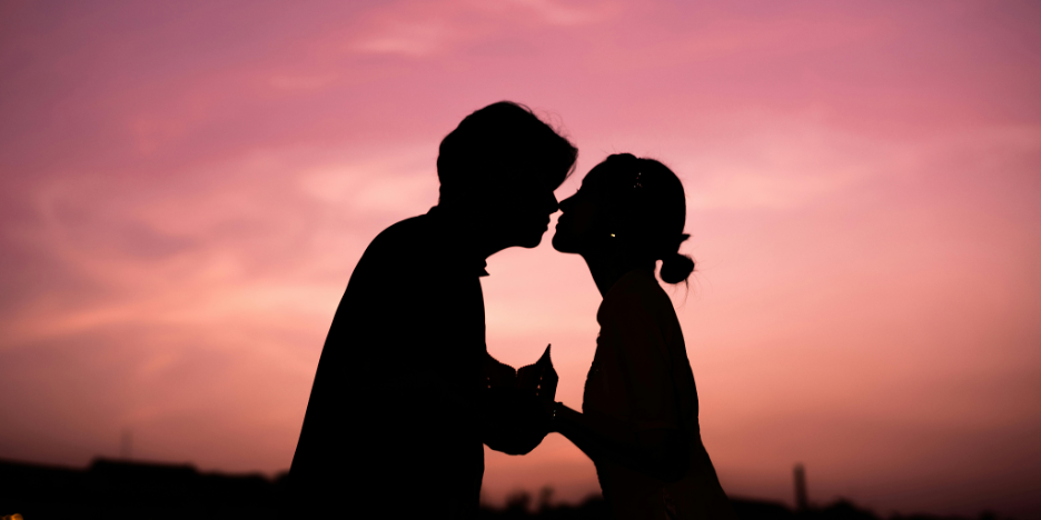 Silhouette of two people leaning in for a kiss with a pink and orange sky behind them.
